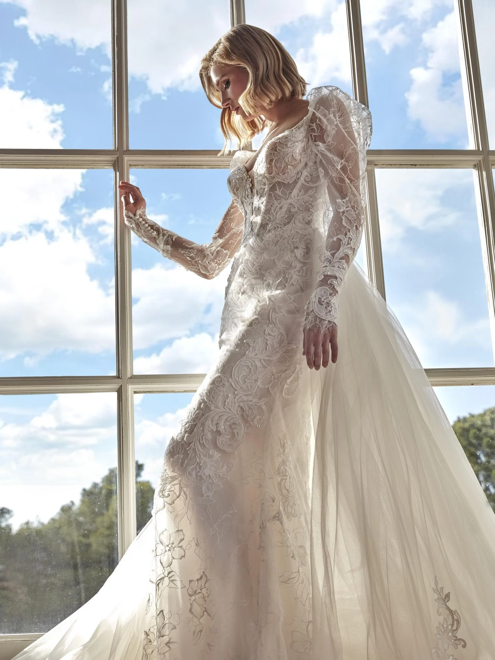 Robe de mariée Biennia – Raffinement romantique et manches longues en dentelle Laissez-vous séduire par l’élégance intemporelle de Biennia, une robe de mariée à manches longues conçue pour les mariées à la recherche d’un style à la fois royal et moderne. Avec sa silhouette sirène délicatement structurée et ses détails floraux brodés, Biennia incarne le romantisme à l’état pur. Le corsage, finement travaillé, présente un décolleté en cœur profond, soutenu par un jeu de transparence subtil et de dentelle illusion. Les manches longues en tulle brodé, légèrement bouffantes aux épaules, apportent une touche de grâce et de noblesse. La jupe sirène est magnifiée par une somptueuse surjupe en tulle brodée, qui ajoute volume et fluidité, tout en créant une traîne majestueuse ornée de motifs floraux raffinés. Biennia marie harmonieusement sensualité, romantisme et tradition pour une allure exceptionnelle.